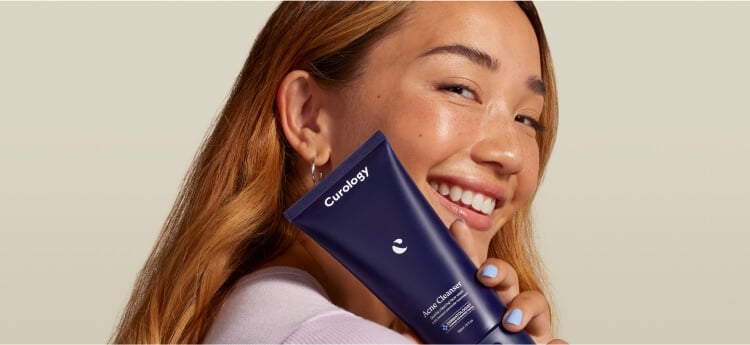 A smiling woman holding a bottle of Curology cleanser next to her face.